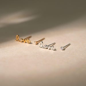 Może przedstawiać: Cztery kolczyki sztyfty w odcieniach złota i srebra. Dwa kolczyki piszą słowo "miłość" kursywą, a dwa pozostałe mają pojedynczy, przezroczysty klejnot. Kolczyki są prezentowane na neutralnej powierzchni.