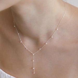 May include: A delicate silver necklace with small, evenly spaced pearls. The necklace has a Y-shaped design, with a strand of pearls cascading down the centre. The necklace is worn around the neck.