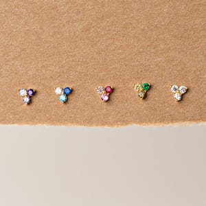 May include: Five pairs of gold stud earrings, each featuring a cluster of three colorful gemstones. The gemstones include purple, blue, pink, green, and clear, set in a triangular arrangement. The earrings are displayed on a neutral, textured background.