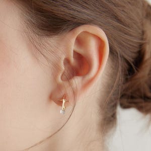 May include: Close-up of a gold-colored star-shaped earring with a small, clear gemstone dangling below. The earring is worn on an ear, with the focus on the jewelry's design and the ear's shape. The earring's design is simple and elegant.
