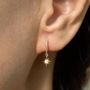 Pode incluir: Um brinco de argola dourado com pequenas pedras transparentes e um pingente de estrela pendurado com uma pedra central semelhante à opala. O brinco é mostrado em uma vista de perto, destacando os detalhes da joia.