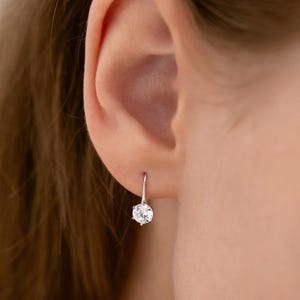 May include: A single silver earring with a large, round, clear gemstone is shown. The earring is dangling from the ear of a person. The gemstone is the focal point, reflecting light and adding sparkle.