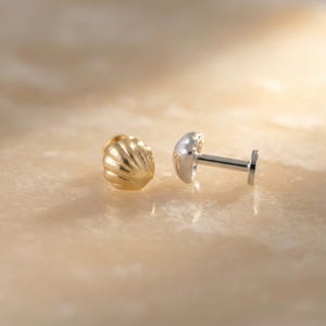 May include: A gold-toned stud earring shaped like a seashell with a ribbed texture, positioned next to a silver-toned flat-back labret post with a smooth, rounded top, showcasing the front and side profiles of the jewelry components.
