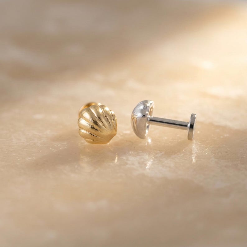 May include: A gold-toned stud earring shaped like a seashell with a ribbed texture, positioned next to a silver-toned flat-back labret post with a smooth, rounded top, showcasing the front and side profiles of the jewelry components.