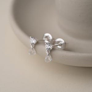 Puede incluir: Un par de pendientes de plata con una piedra transparente y brillante de talla redonda en la parte superior y una piedra en forma de lágrima colgando. Los pendientes se muestran sobre una superficie curva de tono neutro.