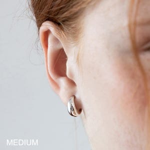 May include: A close-up of a silver hoop earring. The earring is a medium size and is worn on an ear. The word "MEDIUM" is printed in white at the bottom of the image.