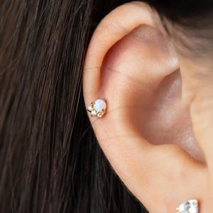 May include: A gold-colored earring with a cluster design. The earring features a central, round, white opal stone, accented by smaller clear crystals. The earring is set against the skin of a person's ear.
