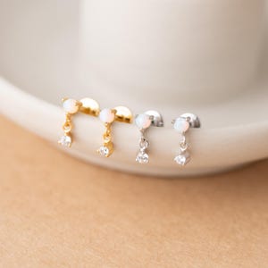 May include: Close-up of four stud earrings. Two are gold-toned, and two are silver-toned. Each earring features a small, round, white opal stone at the top, with a dangling clear cubic zirconia stone. The earrings are displayed on a white surface.
