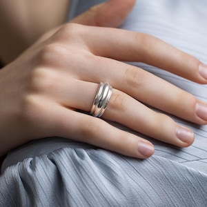 May include: A polished silver ring featuring a triple-band design with smooth, rounded edges. The ring consists of three interconnected bands that create a layered, stacked appearance, reflecting light across its metallic, reflective surface.