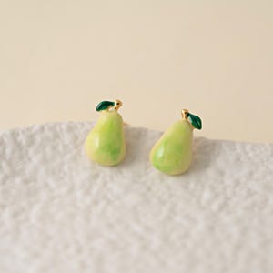以下が含まれることがあります： 洋梨型のスタッドピアス。ピアスは明るい緑色で、濃い緑色の葉と金色のアクセントが付いています。ピアスは白いテクスチャのある表面にあります。