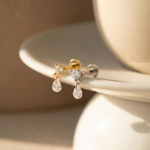 May include: Two stud earrings with flat back posts, one in gold tone and one in silver tone. Each earring features a round clear stone at the post and a dangling pear-shaped clear stone drop. The metal components are polished and reflective.