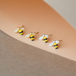 May include: Four pairs of bee-shaped stud earrings. Two pairs are gold-toned, and two are silver-toned. Each bee has a yellow and black striped body, with crystal-like wings. The earrings are displayed on a peach and brown surface.
