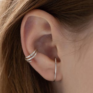 Puede incluir: Primer plano de una oreja adornada con joyas de plata. Un brazalete de oreja de tres bandas se curva alrededor de la parte superior de la oreja, y un pendiente de aro fino de plata cuelga del lóbulo. Las joyas contrastan con el tono de la piel y el cabello castaño.