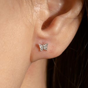 May include: A small silver butterfly stud earring featuring clear, sparkling stones embedded in the wings. The butterfly shape is detailed with a central line and symmetrical wings, providing a delicate and polished appearance on the earlobe.