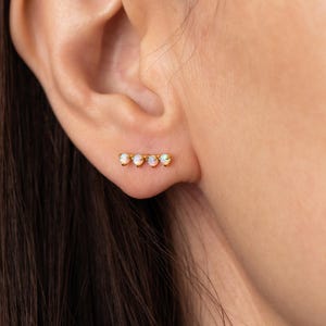 May include: A close-up of a gold bar earring with four small, oval-shaped opals. The earrings are set in a gold metal and are worn on the ear. The background is a blurred image of a person's ear and skin.