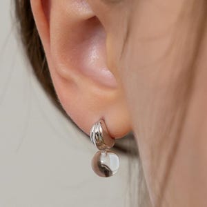 May include: A silver earring with a rounded top and a clear, spherical bottom. The top portion has a ridged design. The earring is shown on a person's ear.