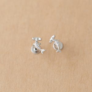 Könnte beinhalten: Ein Paar silberfarbene Ohrstecker. Jeder Ohrring ist wie ein Hammerhai geformt, mit einem kleinen, runden Stecker-Verschluss. Der Körper des Hais ist mit kleinen, klaren Kristallen verziert. Die Ohrringe sind vor einem beigefarbenen Hintergrund.