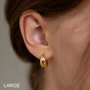 May include: A close-up of a gold-colored hoop earring. The earring has a smooth, polished surface and a teardrop shape. The word "LARGE" is printed in white at the bottom of the image. The earring is worn on an ear.