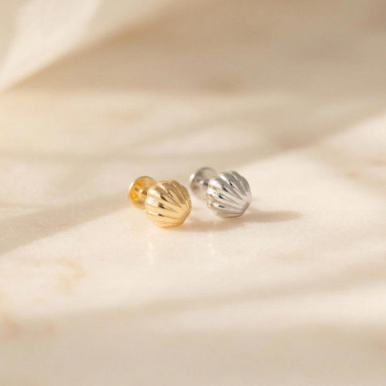 May include: Two small stud earrings shaped like seashells, one finished in a polished gold tone and the other in a polished silver tone. Each earring features a rounded, fan-like design with vertical ridges radiating from the base to the edge.