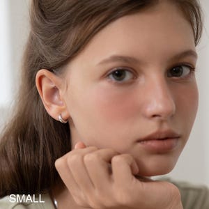 May include: A close-up of a person wearing a small, silver hoop earring. The earring is a simple, rounded design. The word "SMALL" is visible at the bottom of the image.