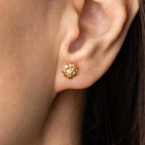 May include: A gold-colored daisy stud earring with a small, clear gemstone in the center. The earring is worn on an ear, showcasing its delicate floral design and sparkling accent. The earring is a piece of jewelry.