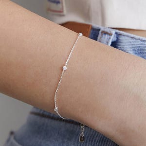 Puede incluir: Una delicada pulsera de cadena plateada con tres pequeñas piedras redondas y transparentes engastadas a intervalos a lo largo de la cadena. La pulsera tiene un diseño de eslabones de metal pulido y un pequeño dije colgante en el cierre.