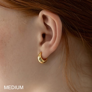 May include: Close-up of a gold-colored, medium-sized hoop earring. The earring is a smooth, curved design, reflecting light. The word "MEDIUM" is printed in white at the bottom of the image. The earring is worn on an ear.