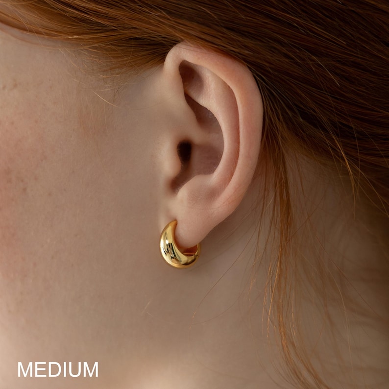 May include: Close-up of a gold-colored, medium-sized hoop earring. The earring is a smooth, curved design, reflecting light. The word "MEDIUM" is printed in white at the bottom of the image. The earring is worn on an ear.