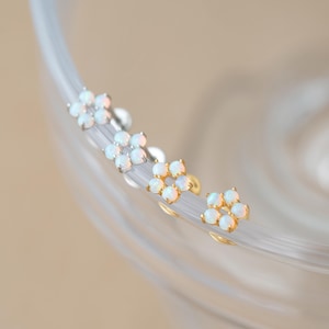 May include: Four flower-shaped stud earrings arranged in a row. Each earring features five small, round, iridescent white opal cabochons set in a circular pattern. Two earrings have a silver-toned metal setting, and two have a gold-toned metal setting.