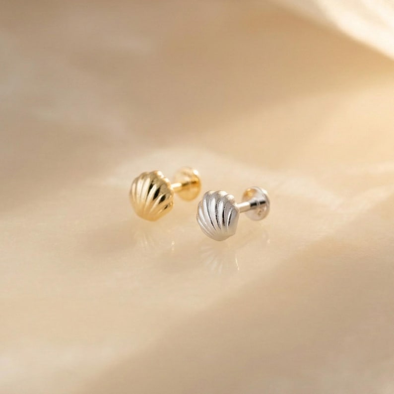 May include: Two stud earrings shaped like small seashells, one finished in a polished gold tone and the other in a polished silver tone. Each earring features a rounded, fan-like design with vertical ridges radiating from the base to the edge.
