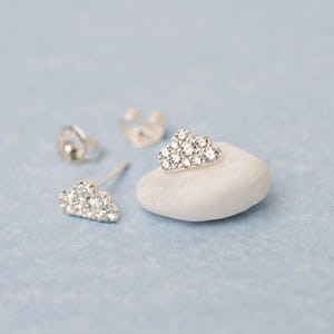 May include: Cloud-shaped stud earrings with sparkling clear stones. The earrings are displayed on a light blue surface with a white stone and earring backs. The earrings are a delicate, feminine accessory.