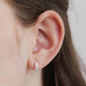 Könnte beinhalten: Nahaufnahme eines silbernen Creolen-Ohrrings mit strukturiertem Design und kleinen, funkelnden Verzierungen. Der Ohrring wird an einem Ohr getragen und zeigt seine komplizierten Details und seinen eleganten Stil.