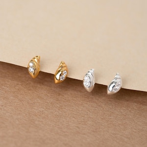 May include: Two pairs of stud earrings, one pair in gold and one pair in silver. Each earring features a curved, shell-like shape with two small, clear, round-cut stones embedded in the centre. The earrings are arranged in a row on a flat surface.