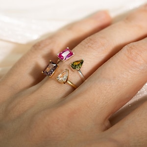 Peut inclure: Un ensemble de deux bagues ouvertes ornées de pierres précieuses multicolores. Une bague présente une pierre rose rectangulaire et une pierre violette rectangulaire. L'autre bague comporte une pierre claire et une pierre vert olive en forme de poire.