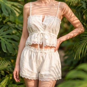May include: A white two-piece set consisting of a sleeveless crop top and matching shorts. The top features thin shoulder straps, a front button placket, a tie-front detail, and delicate lace trim. The shorts have an elasticized waist and lace trim.