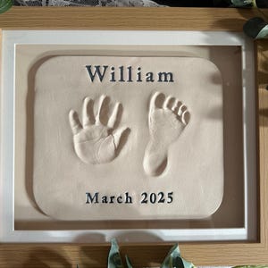 May include: A framed baby handprint and footprint keepsake. The off-white rectangular plaque features an imprint of a hand and foot, with the name "William" and the date "March 2025" in dark lettering. The frame is light brown.