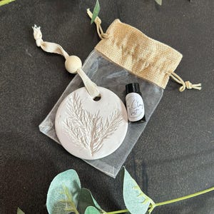 May include: A white, circular clay diffuser with an embossed botanical design, packaged with a small bottle and a burlap bag. The diffuser hangs from a white ribbon with a wooden bead. The bottle has the text "The Clay Nest".