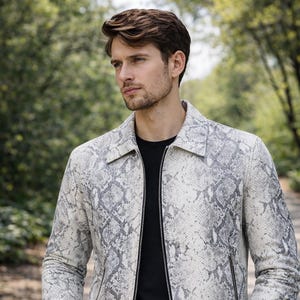 May include: A light-colored jacket with a snakeskin pattern, featuring shades of gray and white. The jacket has a zipper closure and a collared neckline. The person in the image is wearing a black t-shirt and dark jeans.