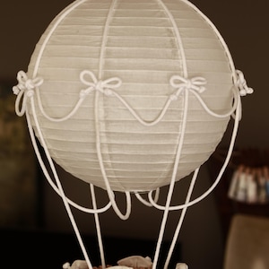 Puede incluir: Una linterna decorativa de papel con forma de globo aerostático. La esfera de papel blanco está adornada con detalles de cuerda blanca y pequeños lazos. Una cesta tejida en la parte inferior contiene pequeños sacos de tela. La linterna cuelga de un solo cordón blanco.