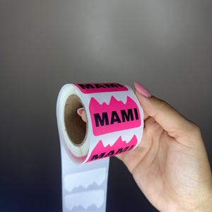 Puede incluir: Un rollo de pegatinas rectangulares con fondo rosa brillante, borde superior en forma de montaña y la palabra MAMI impresa en letras mayúsculas negras en negrita. Las pegatinas están dispuestas en una tira vertical sobre papel blanco.