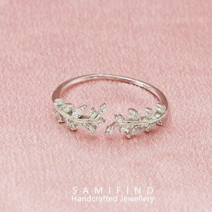Può includere: Un anello d'argento con un design a foglie. L'anello presenta due rami di foglie aperti adornati con piccole gemme trasparenti. L'anello è posto su uno sfondo rosa. Il testo "SAMIFIND Handcrafted Jewellery" è in basso.