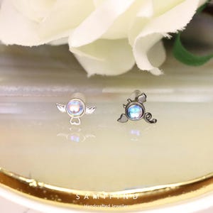 May include: Two small, silver earrings with iridescent, round centre stones. One earring features a wing design, the other a stylized, swirling shape. The earrings are displayed on a light-coloured surface with the text "SAMIFIND Handcrafted Jeweller".