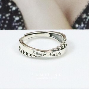 以下が含まれることがあります： 「keep Smile」の文字が刻まれたシルバーリング。リングの片側には編み込み模様、もう片側にはテクスチャパターンがあります。リングは調整可能です。