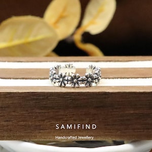 Puede incluir: Un anillo plateado con un diseño floral. El anillo está adornado con pequeñas flores detalladas en forma de margarita. El anillo se muestra sobre una superficie de madera con el texto "SAMIFIND Handcrafted Jewellery".