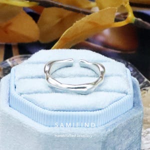 Puede incluir: Anillo plateado, abierto, con un diseño ondulado y orgánico. Se muestra sobre una caja de joyería de terciopelo azul claro. El texto "SAMIFIND Handcrafted Jewellery" es visible en la parte inferior de la imagen.