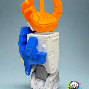 May include: A 3D-printed robot figure with an orange hand, blue arm, and white body. The figure has a geometric design and is set against a light blue background. The figure has the text "MONOKREEPS" on the bottom right.