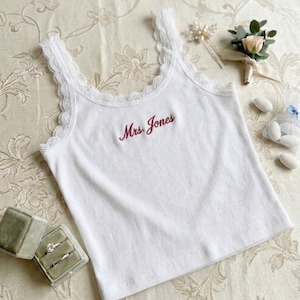Puede incluir: Camiseta sin mangas blanca acanalada con ribete de encaje en las correas. Las palabras "Mrs. Jones" están bordadas en escritura burdeos. Anillos de boda en una caja de terciopelo verde son visibles.