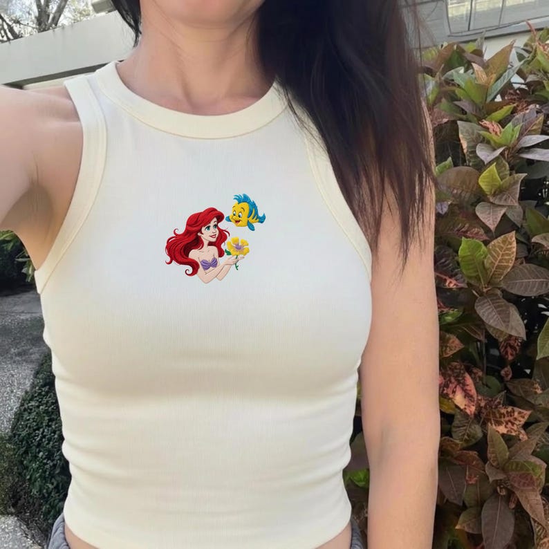Puede incluir: Camiseta sin mangas color crema con un gr&aacute;fico colorido de Ariel, la Sirenita, sosteniendo una flor amarilla, con Flounder. La camiseta sin mangas tiene un cuello redondo y est&aacute; hecha de una tela suave y acanalada.