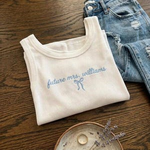 May include: White ribbed tank top with the embroidered text "future mrs. williams" in light blue, along with a bow graphic. The tank top is paired with distressed blue jeans, a gold ring, and a small decorative bowl.