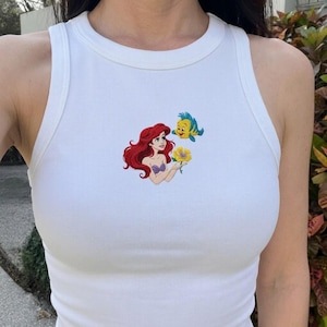 Puede incluir: Camiseta sin mangas blanca acanalada con un dise&ntilde;o bordado de Ariel, una sirena pelirroja, sosteniendo una flor amarilla, con un pez amarillo y azul. La camiseta sin mangas tiene un cuello redondo y un dise&ntilde;o sin mangas.